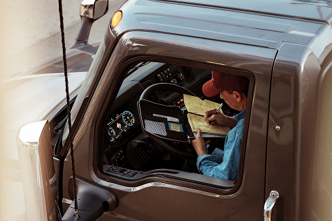 Conductor dentro de un camión INTERNATIONAL gris anotando en una libreta apoyada sobre el volante, con el tablero iluminado al fondo